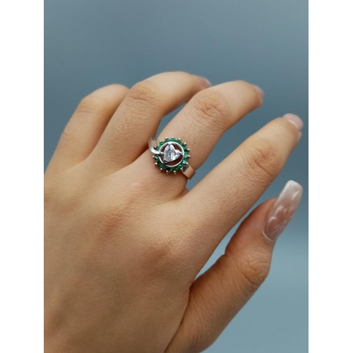 110 - A PLATINUM RING CONTAINING A HEART SHAPED CENTRAL DIAMOND SURROUNDED BY EMERALDS.
6.8gms     SIZE K/... 