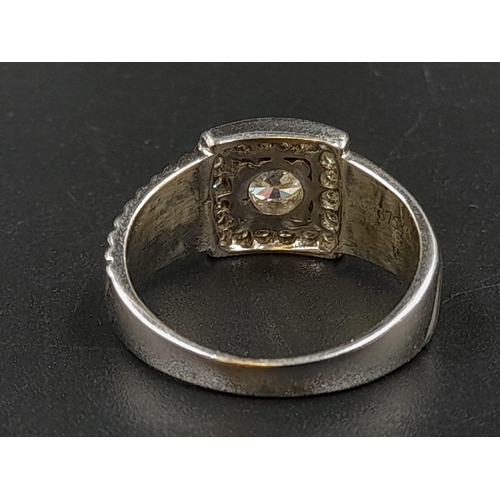 1370 - A 9K White Gold Belt-Buckle Diamond Ring. Central stone surrounded by a square belt-buckle of diamon... 