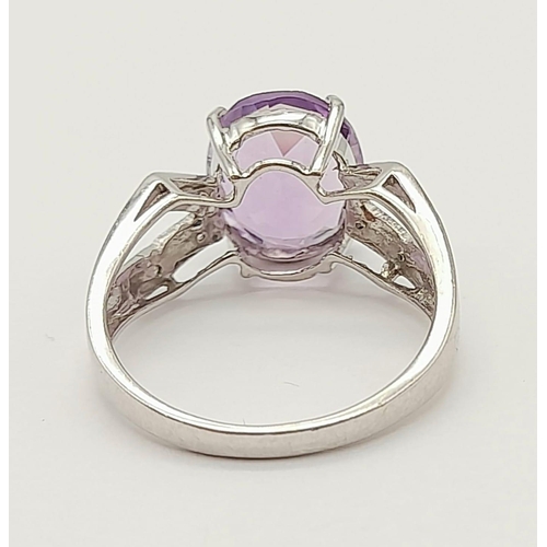 1438 - A 9K White Gold Amethyst and Diamond Ring. Central clean oval amethyst with wings of diamond decorat... 