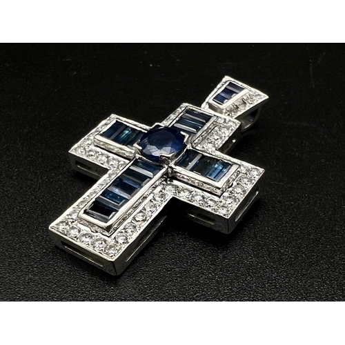 1083 - An 18K White Gold and Sapphire Cross Pendant. A central round-cut blue sapphire is complimented with... 