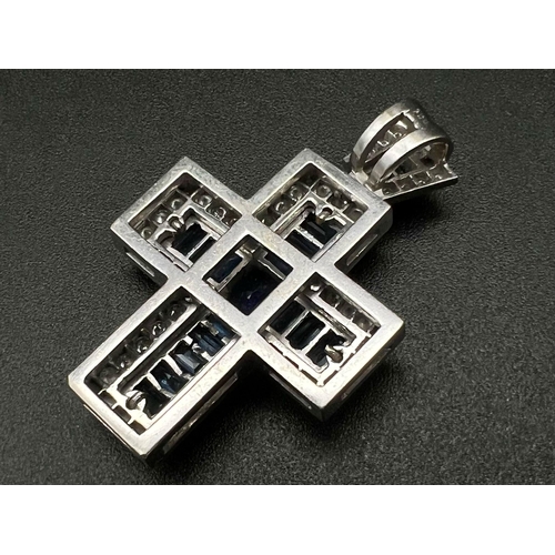 1083 - An 18K White Gold and Sapphire Cross Pendant. A central round-cut blue sapphire is complimented with... 