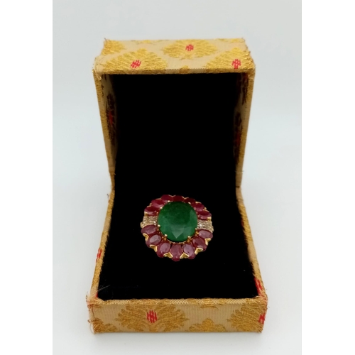 898 - An Emerald & Ruby Gilded Ring. Oval cut. Oval cut emerald with a halo of rubies with diamond accents... 