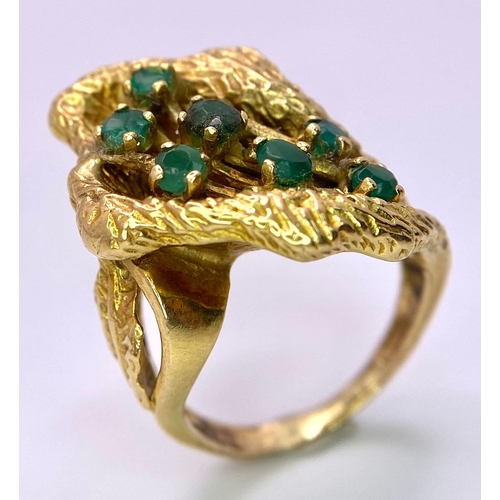 100 - A Vintage 18K Gold (tested) and Emerald Forest Ring. Seven green emerald blossoms sit amongst decora... 