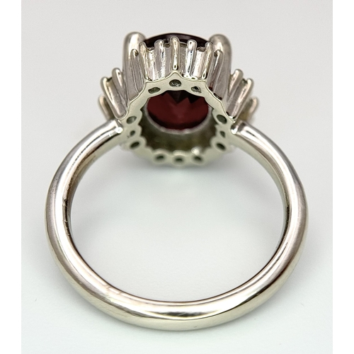 384 - A 14K White Gold, Garnet and Diamond Ring. Rich, oval cut central garnet with a twelve stone diamond... 