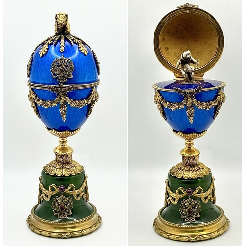 A Diamond Set Gilded Silver, Enamel and Nephrite Jade Surprise Egg. Russian Imperial style 'Easter Egg' with ovoid silver-gilt mounted body in translucent blue. A hinged cover with an elephant finial over a diamond encrusted star, and diamond and ruby coloured stone set twin headed bird motifs. The lower section is decorated with floral garlands. Nephrite jade plinth base with a diamond set twin headed bird motif on a circular foot. Opens to a silver bear with gem set eyes playing a drum on a fine guilloche enamel blue base. Comes with a wooden display case. 18cm tall. 310g total weight.