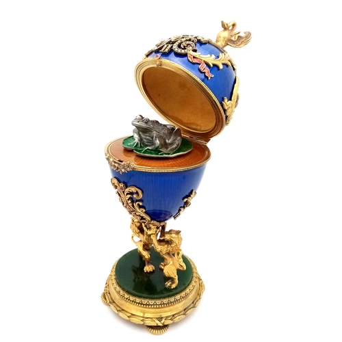 A Russian Diamond Set Gilded Silver, Enamel and Nephrite Jade Surprise Egg. Russian Imperial style 'Easter Egg' with ovoid silver-gilt mounted body in translucent blue. A hinged cover with a swan finial over a pierced scrolling band of diamonds, a diamond set cypher for Nicholas II flanked by red enamel scrolls, and silver-gilt crowns within laurel wreaths. Raised on a base modelled as a pair of lions over a circular nephrite jade base on four feet. Opens to a naturalistic silver frog with gem set eyes raised on a green enamelled lily pad on a yellow guilloche enamel base. Comes with a wooden display case. 18cm tall. 338.2g total weight. UK Mainland Shipping Only or arrange a courier service.