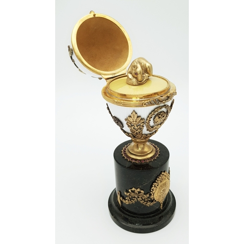 A Russian Style Diamond Set Gilded Silver and Enamel Surprise Egg. Russian Imperial style &lsquo;Easter Egg&rsquo; with ovoid silver-gilt mounted body in white guilloche enamel, decorated with a diamond set 1913 motif, imperial eagle mounts, ribbon and laurel wreath detailing, and red stone accents. Opens to reveal a naturalistic silver mouse figure with purple gem set eyes. Raised on a black hardstone column base with further gilded decoration. Approx 19.9cm height. Total weight 580.7g. Comes with a presentation box. UK Mainland Shipping Only or arrange a courier service.