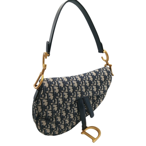 A Christian Dior Oblique Saddle Bag. Navy blue and beige Dior Oblique monogram canvas exterior with navy leather trim, gold-tone hardware, rear open pocket, detachable adjustable leather shoulder strap, signature CD strap attachments and hanging D charm. Saddle-shaped flap opens to a beige textile interior with a zipped inner side pocket. Comes with dust cover bag, authenticity card and certificate of authenticity. Approx. 26cm length x 20cm height. Very light pink mark to the interior of the rear pocket, otherwise overall in good condition. Please see photos or request a condition report. Ref: 29817.