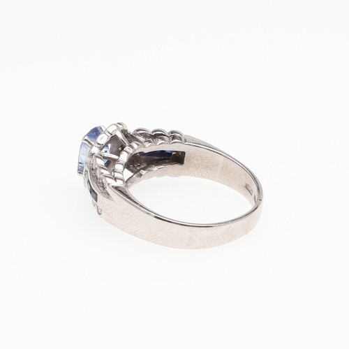 1128 - A SAPPHIRE AND DIAMOND CLUSTER RING. the oval-shaped sapphire is set within a surround of circular-c... 