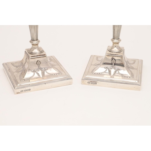 223 - A PAIR OF EDWARDIAN CANDLESTICKS. on square bases (loaded), with swag decoration, reeded borders & d... 