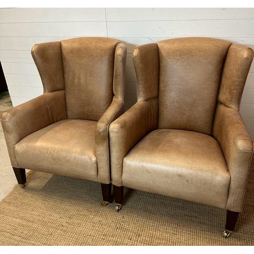 26 - A pair of wing back tan leather arm chairs with brass castors