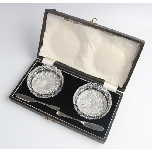 96 - A cased pair of cut glass butter dishes, each 8.7cm wide, each with accompanying silver butter knive... 