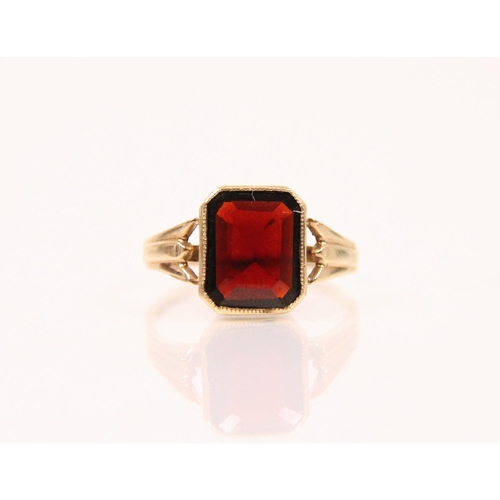 125 - A garnet and 9ct gold ring, the emerald cut garnet within a milgrain setting edge, leading to tapere... 