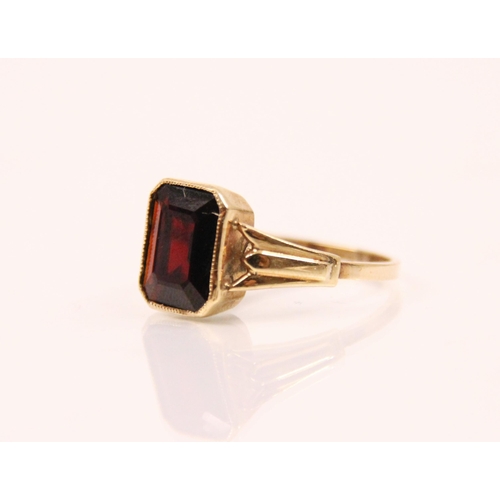 125 - A garnet and 9ct gold ring, the emerald cut garnet within a milgrain setting edge, leading to tapere... 