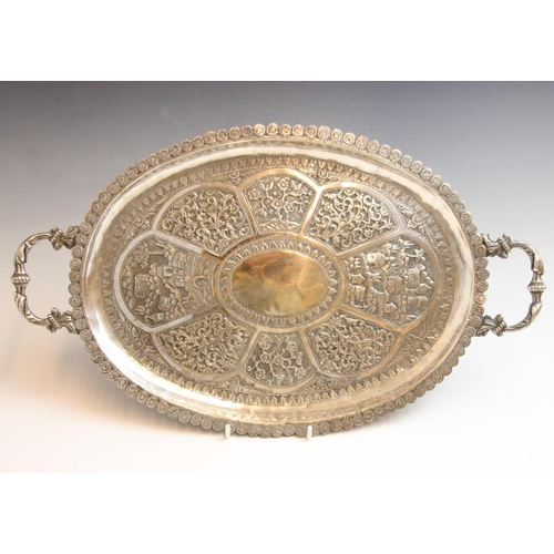 68 - An Indian silver coloured tray, the cast twin handles above oval body decorated in embossed floral a... 