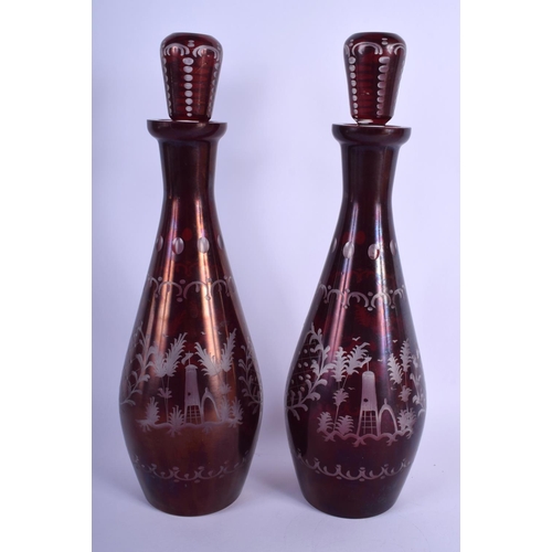 27 - A PAIR OF EARLY 20TH CENTURY BOHEMIAN RUBY GLASS DECANTERS AND STOPPERS decorated with deer and land... 