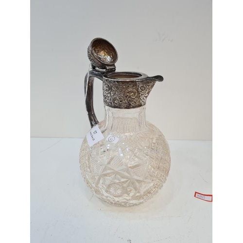 57 - A silver Claret jug having ornate embossed top of foliate scrolls and flowers. Cut glass body of bal... 