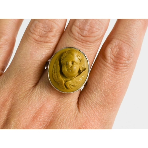 37 - A white metal ring, with relief moulded stone ring in the form of a winged cherub head, size N/O, 6g... 