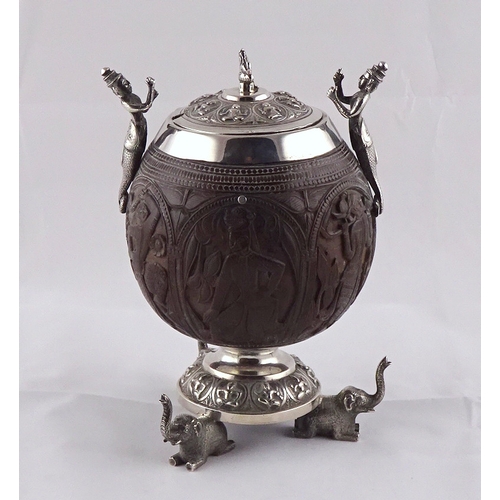 21 - A carved coconut covered pot having white metal handles, mounts and circular stand supported by thre... 