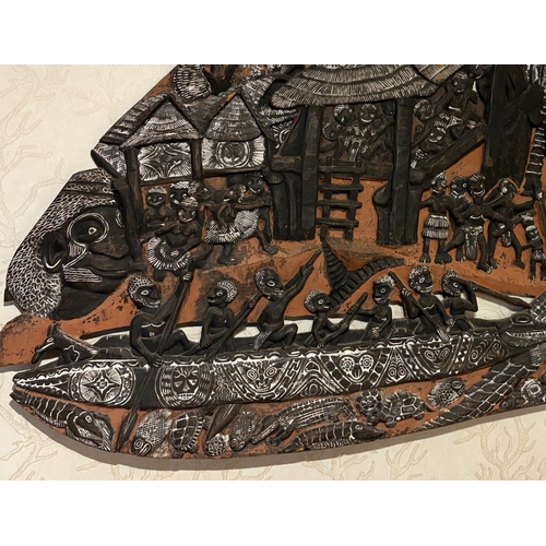 A large Papua New Guinea Storyboard, roughly oval in shape, with scenes from Kambot life in relief. Subject matter of board includes a village and a crocodile hunt below. 150cm x 100cm