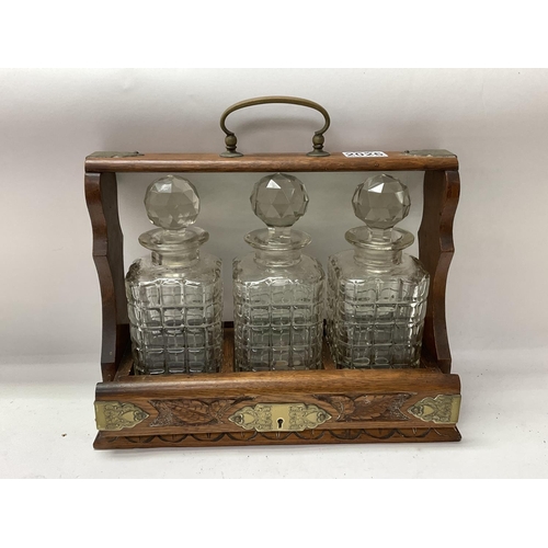 2026 - A carved Oak cased 3 bottle tantalus with engraved brass mount.