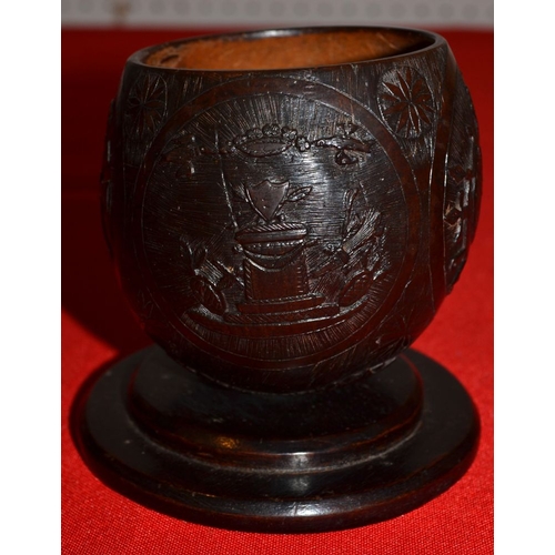 12 - A 19th century coconut cup carved with trophies of music and war on a -platform base 10 cm high