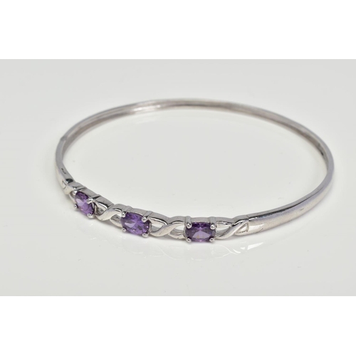 21 - A CUBIC ZIRCONIA BANGLE, designed as three oval purple cubic zirconias interspaced by open cross ove... 
