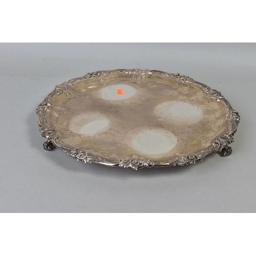 101 - AN EDWARDIAN SILVER CIRCULAR SALVER, cast foliate scroll rim, on three cabriole legs with scroll fee... 