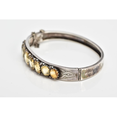 139 - A WHITE METAL CITRINE HINGED BANGLE, designed with a row of graduated claw set, oval cut citrines, o... 