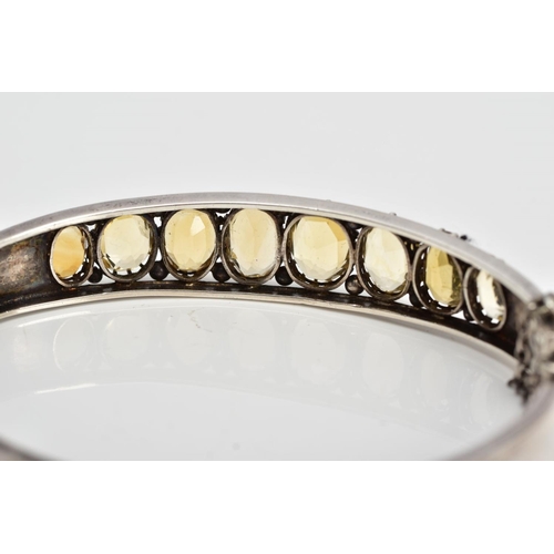 139 - A WHITE METAL CITRINE HINGED BANGLE, designed with a row of graduated claw set, oval cut citrines, o... 