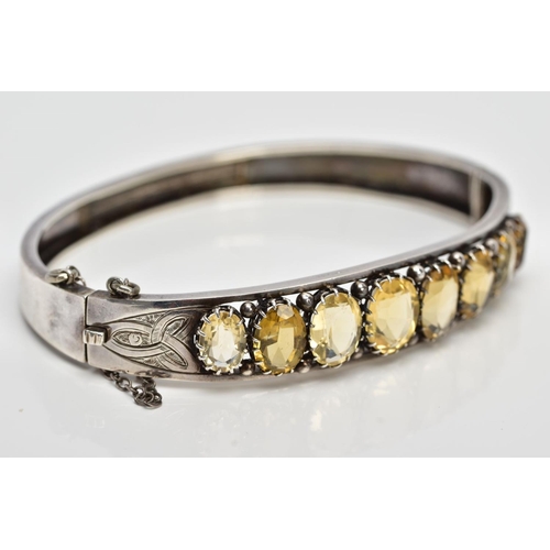 139 - A WHITE METAL CITRINE HINGED BANGLE, designed with a row of graduated claw set, oval cut citrines, o... 