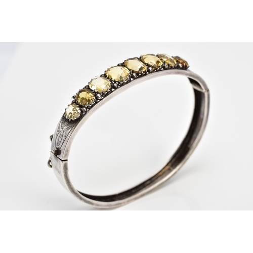 139 - A WHITE METAL CITRINE HINGED BANGLE, designed with a row of graduated claw set, oval cut citrines, o... 