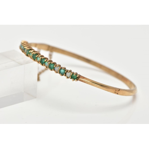 60 - A 9CT GOLD EMERALD AND OPAL BANGLE, hinged bangle, designed with a row of circular cut emeralds inte... 