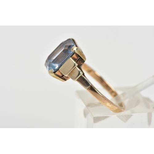 110 - A YELLOW METAL RING, designed with an emerald cut pale blue stone assessed as synthetic spinel, step... 