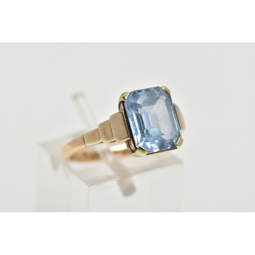 110 - A YELLOW METAL RING, designed with an emerald cut pale blue stone assessed as synthetic spinel, step... 