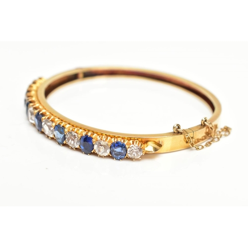 137 - A YELLOW METAL GEMSET BANGLE, yellow metal hinged bangle set with seven oval cut blue sapphires inte... 