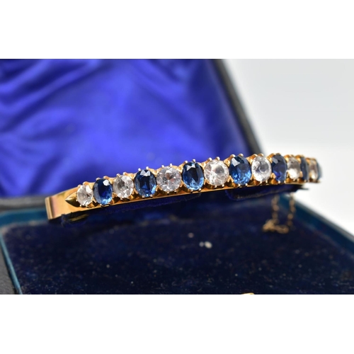 137 - A YELLOW METAL GEMSET BANGLE, yellow metal hinged bangle set with seven oval cut blue sapphires inte... 