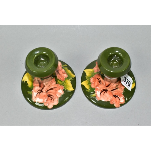 379 - A PAIR OF MOORCROFT POTTERY SQUAT CANDLESTICKS, in Coral Hibiscus pattern on a green ground, impress... 