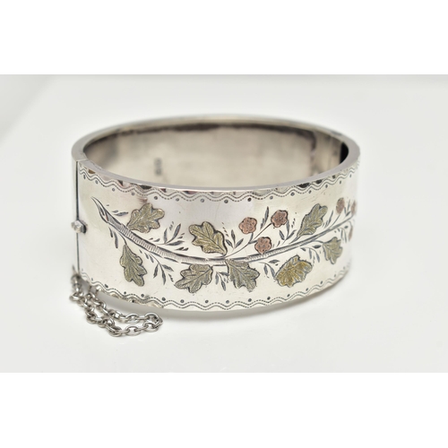 64 - A SILVER HINGED BANGLE,  a wide bangle decorated with a yellow and rose metal foliage etched pattern... 