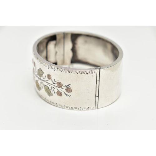 64 - A SILVER HINGED BANGLE,  a wide bangle decorated with a yellow and rose metal foliage etched pattern... 