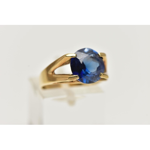 12 - A YELLOW METAL GEM SET RING, a circular cut synthetic blue sapphire, in a four prong setting, bifurc... 