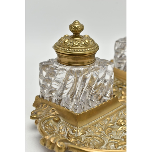 395A - A LATE VICTORIAN BRASS INKSTAND FITTED WITH TWO SQUARE MOULDED GLASS INKWELLS, the stand cast with s... 