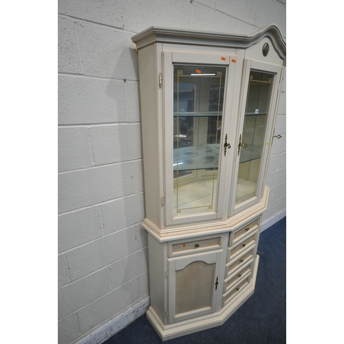 1220 - A 20TH CENTURY CREAM PAINTED CANTED DISPLAY CABINET, the top with an arched crest, three doors enclo... 