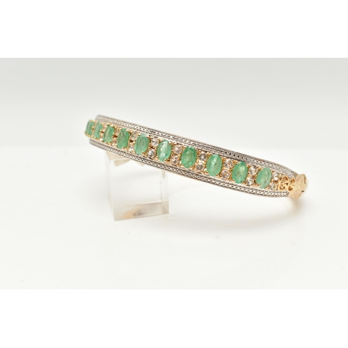 47 - A GILT HINGED BANGLE, designed with a row of eleven oval cut emeralds, each in a four claw setting, ... 