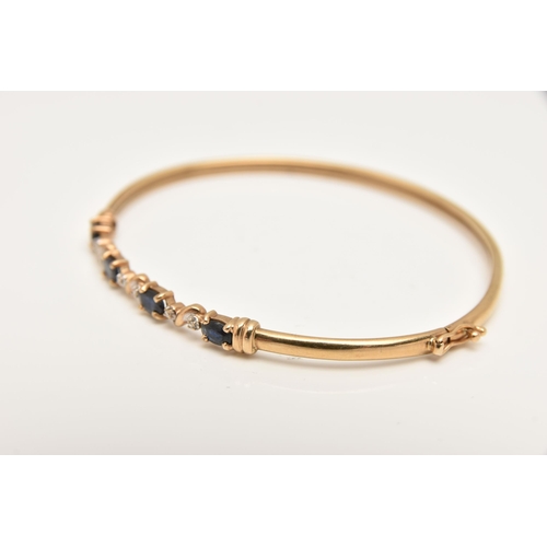 427 - A YELLOW METAL GEM SET BANGLE, set with four oval cut deep blue sapphires interspaced with illusion ... 