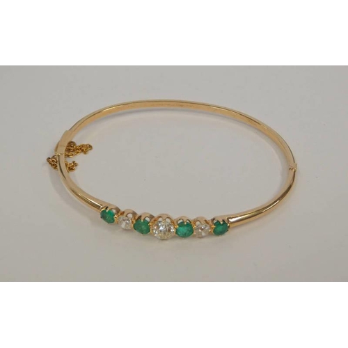 77 - AN EMERALD & DIAMOND YELLOW METAL HINGED BANGLE WITH 4 CIRCULAR CUT EMERALDS AND 3 OLD BRILLIANT CUT... 