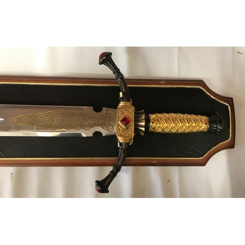 1100 - A large heavy decorative sword on wooden plaque with gold coloured handle and pattern to bottom of b... 