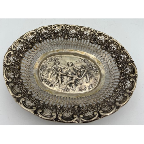 704 - A German silver oval dish depicting dancing cherubs with a pierced border. Size 18 x 14.5cm. Weight ... 