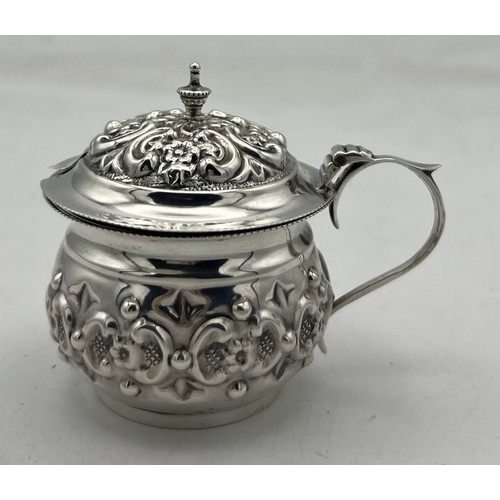 63 - A small bellied silver mustard pot with a repeating pattern of rococo Cs reversed and containing flo... 