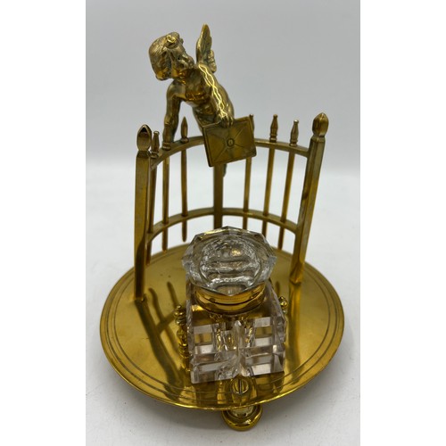 1194 - A 19thC brass inkstand and brass mounted glass inkwell formed as a cherub leaning over a fence envel... 